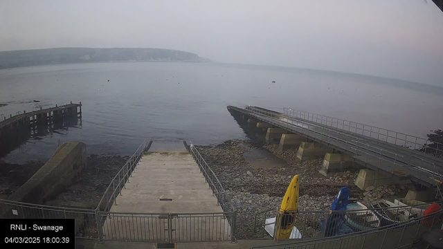 A view of a quiet shoreline with calm water and a misty atmosphere. A wide, concrete ramp leads down to the water, flanked by metal railings. On the right side of the image, there are two kayaks, one yellow and one blue, resting on the ground. In the background, a wooden jetty stretches out into the water, with a rocky shore visible nearby. The distant hills are slightly obscured by fog. The overall scene conveys a serene coastal environment.