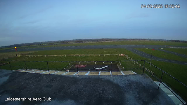 A wide view from a webcam at an aerodrome, showing a runway with a large grassy area surrounding it. In the foreground, there is a black rooftop with a white marking resembling an airplane with an "X" shape. A wooden fence runs along the edge of the green field, and a red light is visible on a pole to the left. The sky is clear with a few clouds, and the image is timestamped.