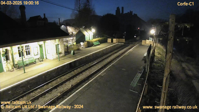 A dimly lit train station at dusk, featuring a stone and wooden structure with a green roof. To the left, there is a covered waiting area with green benches. The platform is empty, with two railway tracks running parallel, leading into the distance. A wooden fence separates the platform from an area with greenery. In the background, a silhouette of a hill with a castle-like structure is visible against the darkening sky. Street lamps provide soft illumination, creating a quiet evening atmosphere.