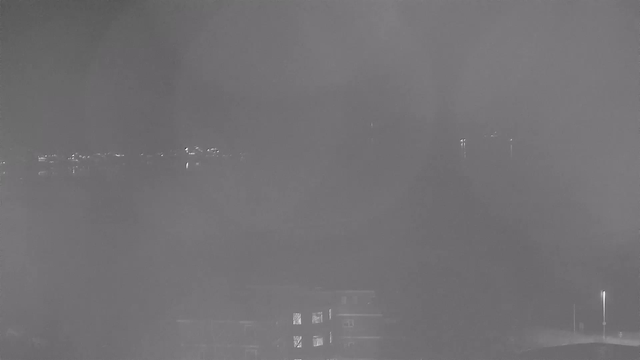 A dimly lit scene captured by a webcam during nighttime. In the background, there are faint lights originating from a distant shore, reflecting on the water, creating a hazy and blurry effect. The foreground shows the outline of a building and some trees, but details are obscured by darkness and fog.