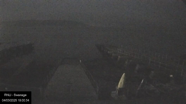 A dimly lit image depicting a harbor scene at night. In the foreground, a wide walkway leads towards a pier, partially obscured by shadow. Several boats are moored nearby, with their outlines faintly visible against the dark water. The horizon blends into the night sky, creating an overall sense of calmness and tranquility despite the low visibility. The date and time are displayed in the bottom left corner.