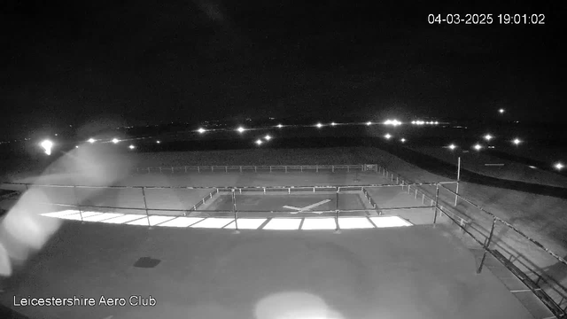 A dark scene captured from a webcam at night. The foreground features a flat rooftop surface with a white illuminated strip near the center, which appears to be part of a structure. There is a railing visible along the edge of the roof, and a pattern resembling an "X" in the center of the illuminated area. In the background, faint lights are scattered in the distance, likely indicating nearby structures or vehicles. The overall lighting is low, with the illuminated areas contrasting against the dark surroundings.