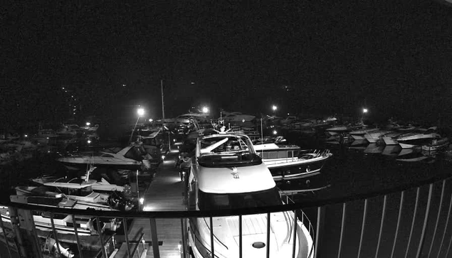 A nighttime view of a marina filled with various boats, illuminated by artificial lights. In the foreground, a large white boat is docked, and several smaller vessels are moored along the sides. The surrounding area is dark, with only the lights from the boats and a few distant buildings providing visibility. The scene has a calm and quiet atmosphere, indicative of a peaceful night by the water.