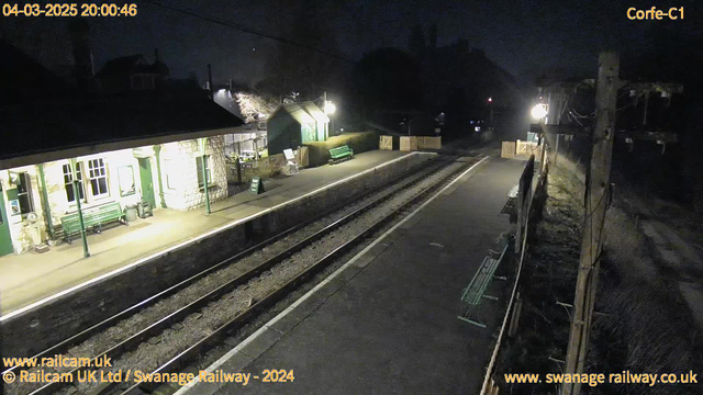 A dimly lit railway station at night with a stone building on the left. The building has large windows and green trim, with benches situated on the platform. To the right, there are wooden barriers and more benches along the platform. A sign indicates "WAY OUT." In the background, faint light silhouettes trees and structures, including a pole with overhead wires. The ground is dark, with visible railway tracks leading into the distance.