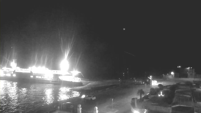 A dark nighttime scene featuring a harbor. In the foreground, a portion of a parking area is visible with parked cars. To the left, a large ferry boat is illuminated by bright lights, reflecting off the water. The background shows faint outlines of structures and additional lights from the shore. The overall atmosphere is calm with a clear sky.