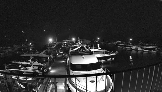 A dimly lit marina at night, featuring numerous boats and yachts docked. The foreground includes a large white yacht, with lights illuminating parts of the surrounding boats. Several smaller vessels are visible in the background, also lit by soft lights. The scene is predominantly dark with contrasting shadows and reflections on the water.