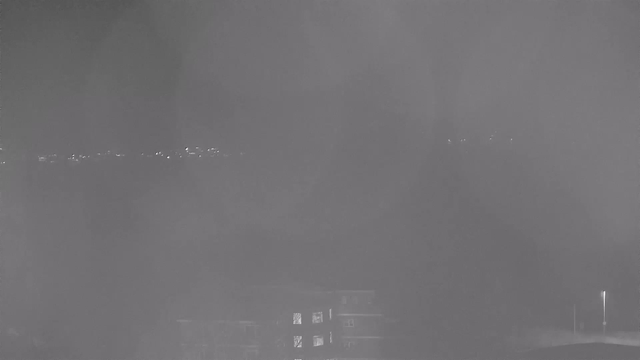 A dimly lit scene captured by a webcam, showing a faint outline of a building at the bottom left with a few illuminated windows. In the background, a distant line of lights suggests a city or town, but details are obscured by low visibility, creating a hazy atmosphere. The overall tone is monochromatic with shades of gray, contributing to the unclear view.