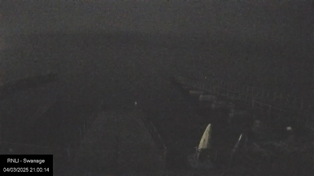 A dark webcam image shows a coastal scene at night. In the foreground, there are vague outlines of boats, with one boat featuring a white sail. The background is largely indistinct, blending into the dark sea and sky, suggesting an overcast or moonless night. The timestamp indicates the image was captured at 9:00 PM.