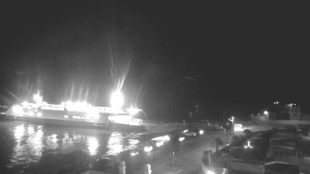 A large ferry is docked at a pier at night, illuminated by bright lights. The water is calm and reflects the lights. There are vehicles, possibly cars, approaching the ferry from a nearby road. The scene is predominantly dark with some background lighting from the pier and vehicles.