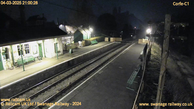 A dimly lit railway station scene shows an empty platform with two metal benches. To the left, there is a stone building with large windows, partly illuminated, and green benches outside. A "Way Out" sign is visible on the ground. The platform is bordered by wooden fencing. The railway tracks run parallel through the scene, and further down the platform, another bench is illuminated. In the background, streetlights cast a soft glow, and a silhouette of trees and buildings is visible against the night sky.