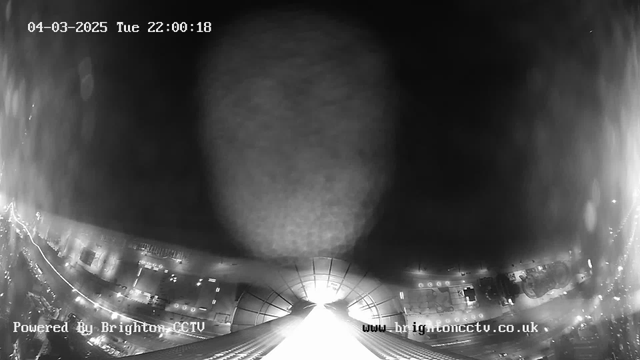 A black and white webcam image taken at night, showing a view upwards from a building. The center of the image features a bright light source, possibly from the building's top, with a circular reflection visible. Below, faint outlines of streets and buildings can be seen, illuminated by streetlights. The date and time displayed are March 4, 2025, at 10:00:18 PM. The bottom left corner has text indicating the image is powered by Brighton CCTV.