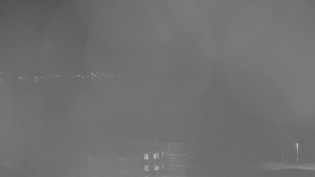 A nighttime webcam image showing a faint outline of buildings with some illuminated windows in the foreground. The background features distant lights, likely from a city or roadway, blurred by condensation or fog, creating a hazy atmosphere.