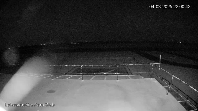 A dark scene captured by a webcam at the Leicestershire Aero Club. The foreground shows a flat surface, likely a runway, with faint markings. A railing runs along the lower edge of the image. In the background, there are distant lights, suggesting an urban area beyond the dark horizon. The overall atmosphere is quiet and nighttime, with minimal visibility. The date and time in the corner indicate March 4, 2025, at 10:00 PM.