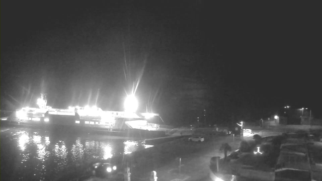 A dark nighttime scene depicting a large ferry docked at a harbor, illuminated by bright lights on the vessel. The water reflects the lights, creating a shimmering effect. In the foreground, there are vehicles parked along the dock, and some pedestrians are walking nearby. The surrounding area is dimly lit with a few additional lights visible in the background.