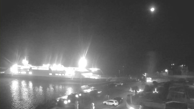 A dark scene featuring a brightly lit ferry docked at a harbor. Reflections of the lights shimmer on the calm water. In the foreground, several vehicles are parked along the harbor, with some visible illumination from streetlights. The moon is visible in the night sky, providing a faint glow. The surroundings are mostly shadowed with only small areas of illumination from lights.