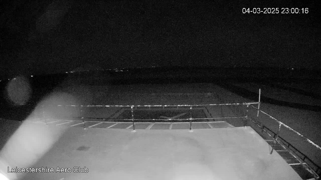 A dark scene captured from a webcam at night, showing a flat surface that appears to be an airfield or runway with marked areas. In the foreground, a railing runs along the edge, suggesting a viewing area or balcony. The background shows faint lights in the distance, indicating a horizon or other structures. The image is black and white, with low visibility overall. The timestamp displays the date and time in the corner.