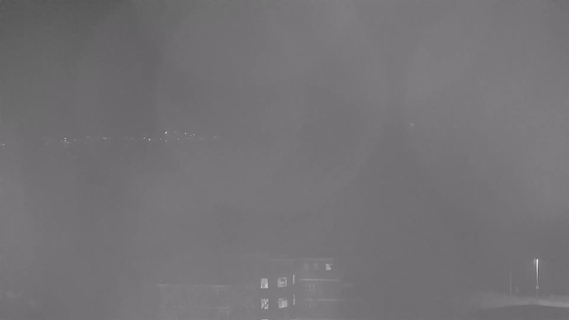 A blurry, monochrome image captured by a webcam, showing indistinct shapes and soft lights. The bottom portion features a dimly lit building with several windows. In the background, faint outlines of distant lights are visible against an unclear sky. Overall, the scene has a foggy or misty appearance, reducing visibility significantly.