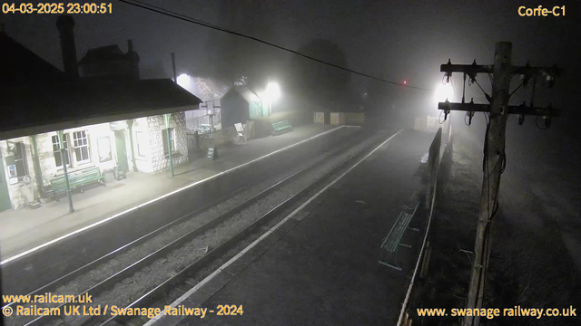 A dimly lit railway station scene shrouded in fog. The foreground shows two sets of railway tracks leading into the distance. On the left, a stone building with windows reflects faint light, while a green bench is situated on the platform. There is a "Park Out" sign visible. A wooden pole with electrical wires stands to the right, where a hazy light source is discernible, suggesting an approaching train. The overall atmosphere is quiet and mysterious, emphasizing the low visibility due to fog.