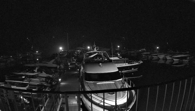 A view of a nighttime marina with various boats docked. The scene is illuminated by several bright lights reflecting off the water. In the foreground, a large white boat is prominently featured, while multiple smaller boats are visible in the background, all resting in their slips. The atmosphere is calm and tranquil, typical of a marina at night.