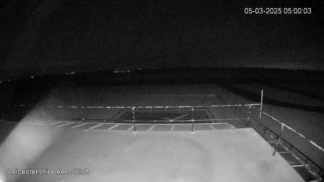 A dark scene showing an empty airstrip at the Leicestershire Aero Club. The image is predominantly black with faint outlines of the runway markings and a fence in the foreground. The timestamp in the corner indicates it is early morning, while distant city lights can be seen on the horizon. The overall atmosphere is dim and quiet, suggesting a stillness typical of early hours.