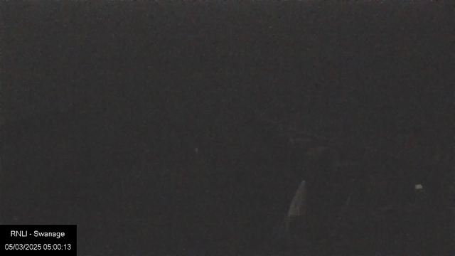 A dark and blurry scene with minimal visible details, suggesting low light conditions. There is a timestamp reading "05/03/2025 05:00:13" located at the bottom left corner, along with the text "RNLI - Swanage." The overall visibility is extremely limited, making it difficult to discern any specific objects or features.