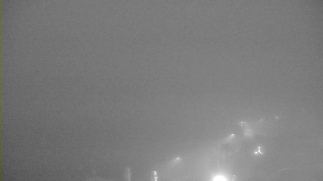 A heavily foggy scene captured by a webcam, with soft lights barely visible in the distance. The image is predominantly gray, creating a dull and muted atmosphere. The outlines of structures are faintly discernible, shrouded in mist, with the overall visibility extremely low.