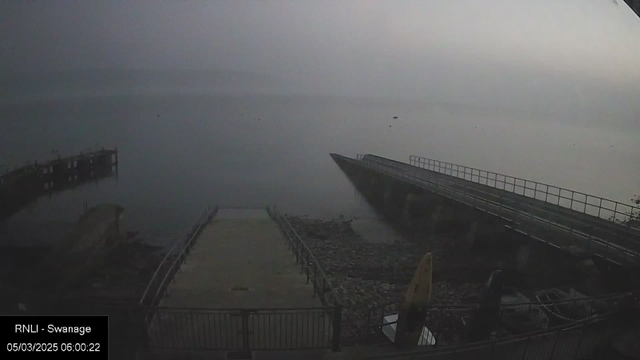 A foggy early morning view of a coastal area. In the foreground, there is a concrete ramp leading down to rocky terrain. To the left, a wooden pier extends into the still water, which reflects the muted colors of the cloudy sky. The scene has a tranquil but misty atmosphere, with little visibility beyond the immediate shoreline. A few boats are partially visible at the bottom right.