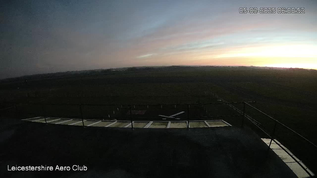 A panoramic view from a webcam at dawn, showing a grassy airfield with a few white markers. In the foreground, there is a flat rooftop with a low railing, and a small aircraft silhouette is visible on the ground. The sky transitions from dark blue to soft pink and orange hues near the horizon, indicating the approaching sunrise. The overall scene conveys a tranquil early morning atmosphere at the Leicestershire Aero Club.