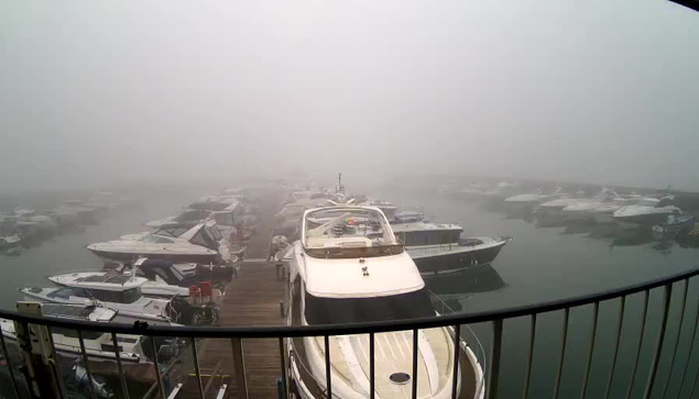 A foggy scene at a marina showing several boats docked along a wooden pier. The visibility is low due to the dense fog, obscuring the background and giving a muted appearance to the scene. The boats vary in size and design, with some being larger motorboats and others smaller vessels. The water reflects the boats and the fog, creating a calm yet indistinct atmosphere. There is a railing in the foreground, indicating the viewpoint from a higher position.