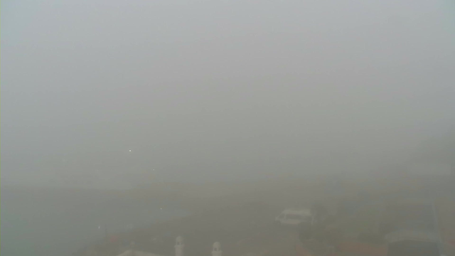 A dense fog covers the area, obscuring visibility almost entirely. The scene is gray and indistinct, with faint outlines of a harbor or coastline potentially visible in the background. There are indistinct shapes that suggest buildings or vehicles, but details are unrecognizable due to the heavy fog. The overall atmosphere is muted and lacking in contrast, creating a sense of an overcast, still environment.