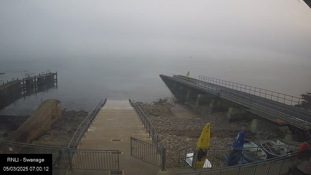 A foggy waterfront scene with a visible stone and concrete pier leading out over calm water. Steps descend from the pier to the shore, where rocks and pebbles are partially exposed. In the distance, a figure in a bright yellow raincoat stands on the pier. Several kayaks in different colors are secured on the shoreline. The atmosphere is gray and muted, contributing to a sense of stillness.