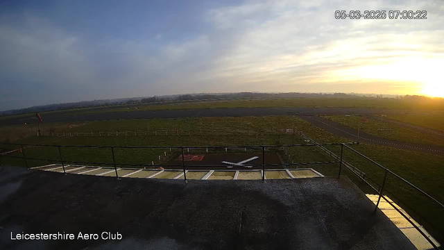 A view from a high vantage point at the Leicestershire Aero Club, capturing the airfield during early morning. The ground is mostly grassy with a few runways visible, and a small aircraft is positioned on a marked area. A railing in the foreground suggests the observation deck, and the sky transitions from pale blue to a warm, golden hue near the horizon as the sun rises.