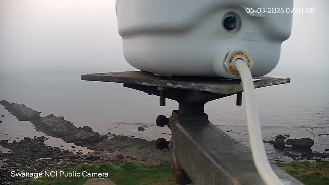 A close-up view of a white cylindrical object mounted on a metallic structure, with a white cable attached. In the background, there is a rocky coastline meeting the water under a cloudy, foggy sky. The image appears to be taken at dawn, with muted colors and low visibility. The date and time are displayed at the top right corner.