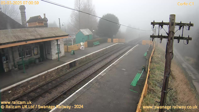 A foggy scene at a train station early in the morning. The view shows empty railway tracks leading into the mist. On the left, there is a stone station building with a sloped roof and several green benches in front. A green shed is visible in the background, along with a wooden fence enclosing some areas. There is a wooden telegraph pole on the right side of the image, with wires extending from it. The overall atmosphere is quiet and subdued due to the fog.