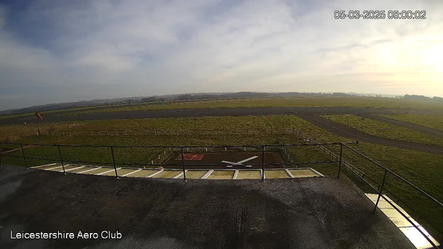 A wide view of an airfield taken from a webcam, with a grassy landscape and a runway in the background. In the foreground, there is a flat rooftop with a railing. A red and white windsock is visible to the left. The sky is partly cloudy with soft light, and the time and date are displayed in the top right corner. In the distance, there are hills and a clear demarcation of the airfield boundaries.