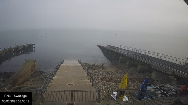 A foggy seascape scene is depicted, with a small concrete ramp leading down to the water in the foreground. On the left, there is a pier extending into the water, while on the right, a metal ramp leads to a wider deck area. Several kayaks in bright colors, including yellow and blue, are positioned near the base of the ramp on stony ground. The water appears calm, but visibility is limited due to the fog, making distant objects indistinct. The time stamp indicates it's early morning.