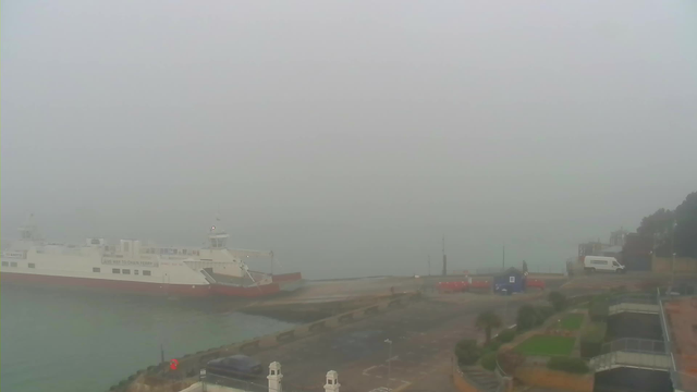 A foggy scene at a harbor. In the foreground, a large white ferry with a red bottom is docked. The ferry is partially obscured by thick fog. There is a parking area with a few vehicles, including a white van, and a small blue building nearby. The water appears calm but is also shrouded in fog, blending seamlessly with the cloudy sky. Surrounding the area are patches of greenery and landscaping. Overall, visibility is very low due to the fog.