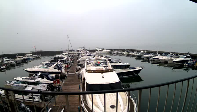 A view of a marina filled with numerous boats of varying sizes docked along wooden piers. The water is calm and reflects the shapes of the boats, while a light fog covers the scene, softening the outlines of the surroundings. In the distance, a few boats are tied to a pier, and there are some flags visible at the edge of the marina. The overall atmosphere is tranquil and somewhat muted due to the fog.