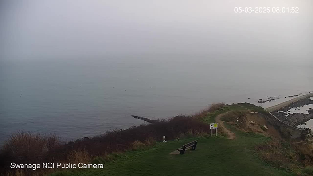 A foggy coastal scene with a calm sea and limited visibility. In the foreground, there is a grassy area with a wooden bench facing the water. To the right, a path leads down the hill toward a rocky shore and a beach. There are some small rocks visible near the water's edge. A sign stands on the grassy area, but it is unclear what it indicates. The time and date in the corner indicate it's early morning.