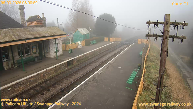 A foggy scene at a train station. The platform is lined with wooden benches and there is a small building with a sloped roof, partially obscured by mist. On the left, a green shed and a sign reading "WAY OUT" are slightly visible. There are two sets of railway tracks that stretch into the fog, and a wooden telephone pole stands on the right side of the image. The atmosphere is quiet and serene, with limited visibility due to the heavy fog.