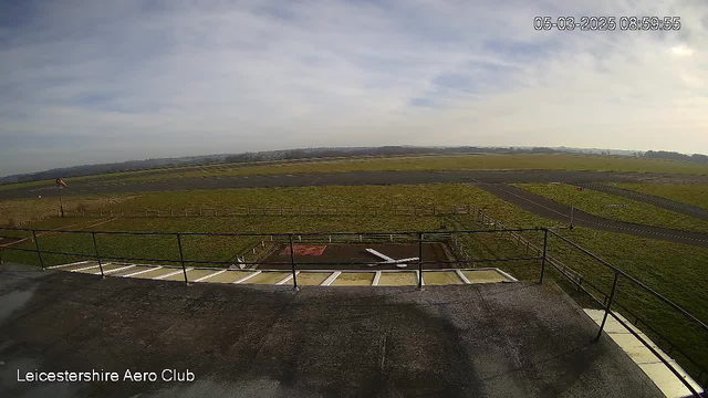 A view from a webcam at the Leicestershire Aero Club. The foreground shows a flat rooftop with a black surface and metal railings. Below, there is a grassy area with a yellowish-green field extending into the background. A small part of a runway, paved with gray asphalt, is visible on the left, while the right side shows a path. The sky is mostly clear with scattered clouds, and the image appears to be taken in daylight. There is a flag at the edge of the grassy area, gently waving in the wind. The timestamp indicates the image was taken in March 2025.