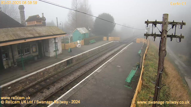 A foggy railway station scene with a platform visible on the left. There are empty green benches along the platform and a few wooden fences in the background. A small building with a sloped roof and a chimney is present, partially obscured by fog. A telephone pole is situated on the right with wires tangled along it. The atmosphere is misty, creating a subdued and indistinct appearance. The ground is made of stone slabs, partially covered by gravel near the train tracks.