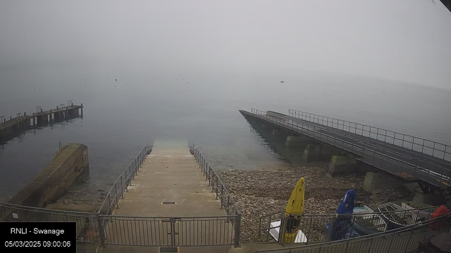 A foggy scene at a coastline. In the foreground, a set of concrete steps leads down to the water, with a railing on either side. To the right, a gently sloping ramp extends towards the water, supported by pillars. On the shore, there are scattered rocks and pebbles. Two canoes are visible, one yellow and one blue, resting on the ground. The water appears still and calm, with limited visibility due to the fog, and a few indistinct shapes, possibly boats, can be seen in the distance.