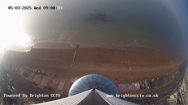 An aerial view of a beach taken from a high vantage point, with the shoreline stretching across the bottom of the image. The water is calm, reflecting sunlight, while a fishing jetty extends into the sea. The beach is sandy, with some structures and people visible. In the foreground, part of a dome-shaped building is visible, suggesting this view is taken from a tall structure. The sky appears bright with some haze. Bottom text indicates the source of the image.