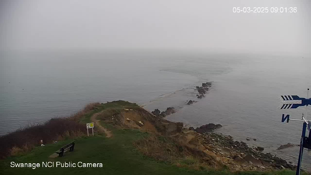 A coastal view shows a gray, overcast sky meeting a calm sea. The shoreline features rocky outcrops extending into the water. There is a grassy cliff in the foreground with a wooden bench facing the sea. A path leads from the bench toward a warning sign near the edge of the cliff. In the distance, the water appears somewhat hazy, creating a blend of sea and sky. A wind direction indicator is visible on the right side of the image.