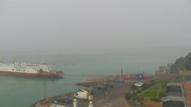 A foggy harbor scene with a white ferry docked at a pier, its ramp extended towards the land. The water is calm and slightly rippled, blending into the gray mist. In the foreground, there is a parking area with several cars, including blue and white vehicles. A yellow truck is parked nearby, under a covered area. To the right, there are steps leading to landscaped gardens with patches of grass and shrubs. The overall atmosphere is muted due to the fog, creating a serene but obscured view.