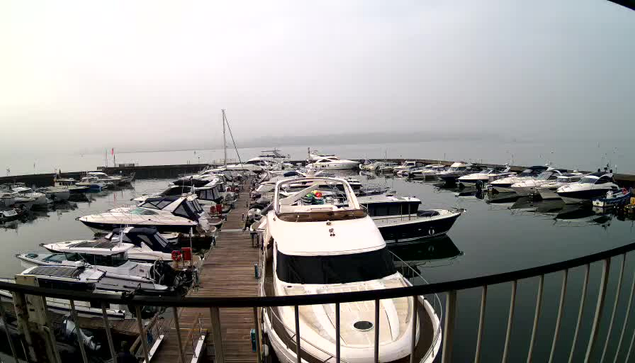 A foggy marina scene featuring numerous boats docked in the water. The foreground shows a wooden walkway leading to several white and blue boats, while additional boats are visible in the background. The sky appears overcast, creating a muted atmosphere, with hints of distant land barely visible through the fog.