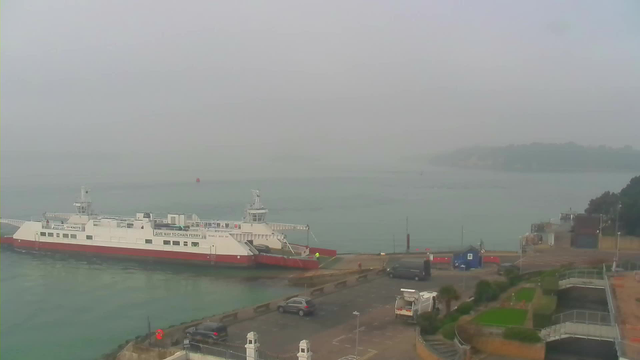 A foggy waterfront scene featuring a large white ferry with red accents docked at a pier. The water is calm and reflects the muted colors of the environment. There are a few vehicles parked on the land, and low buildings can be seen in the background alongside some greenery. The visibility is low due to the fog, obscuring details of the far shore and surroundings.