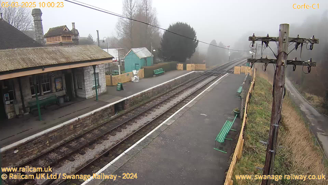 A foggy scene at a railway station. On the left, there is a stone building with a thatched roof, featuring a bench and a poster visible in the entrance. Green benches are lined along the platform. In the background, a wooden fence surrounds a small area with a green shed. The railway tracks run horizontally through the image, leading into the misty distance. A wooden telegraph pole stands on the right, with various wires attached. The atmosphere is quiet and gray due to the fog.