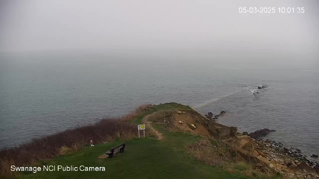 A foggy coastal scene shows a grassy hill with a bench facing the sea. In the background, the water is a muted blue-gray, blending into the misty sky. The coastline features rocky outcrops, and there is sparse vegetation around the bench. A signpost is visible near the bench, indicating a pathway that leads down the hill toward the water. The image is timestamped.